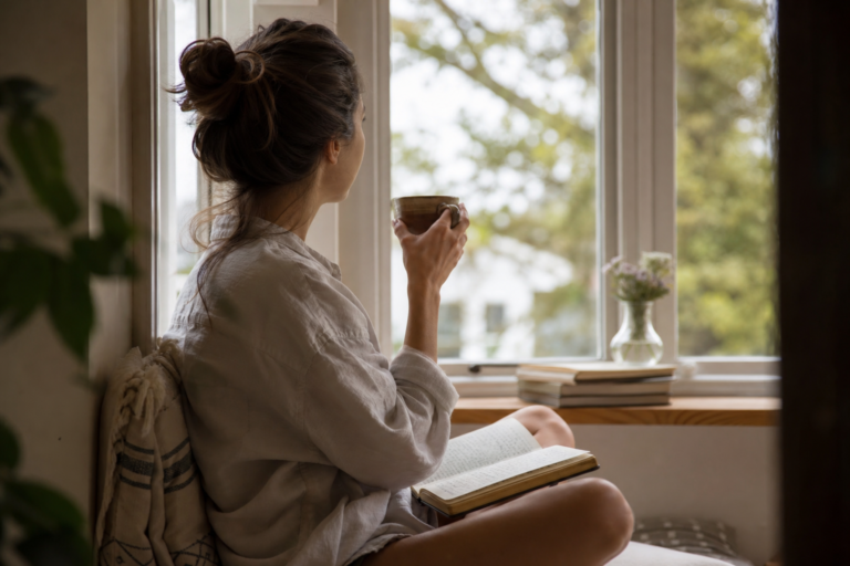 Person sitting by a window with a journal and tea, taking a quiet moment to feel grounded again